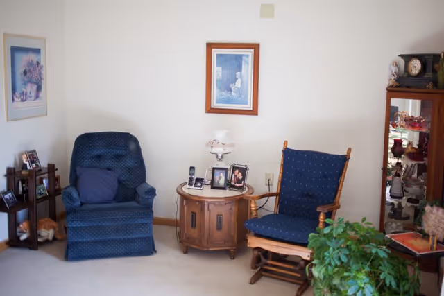 A cozy living room area with two blue upholstered chairs, a wooden side table between them holding framed photos and a lamp, a small wooden shelf with framed pictures and a stuffed animal, a glass display cabinet with decorative items, a green potted plant, and framed artwork on the walls.