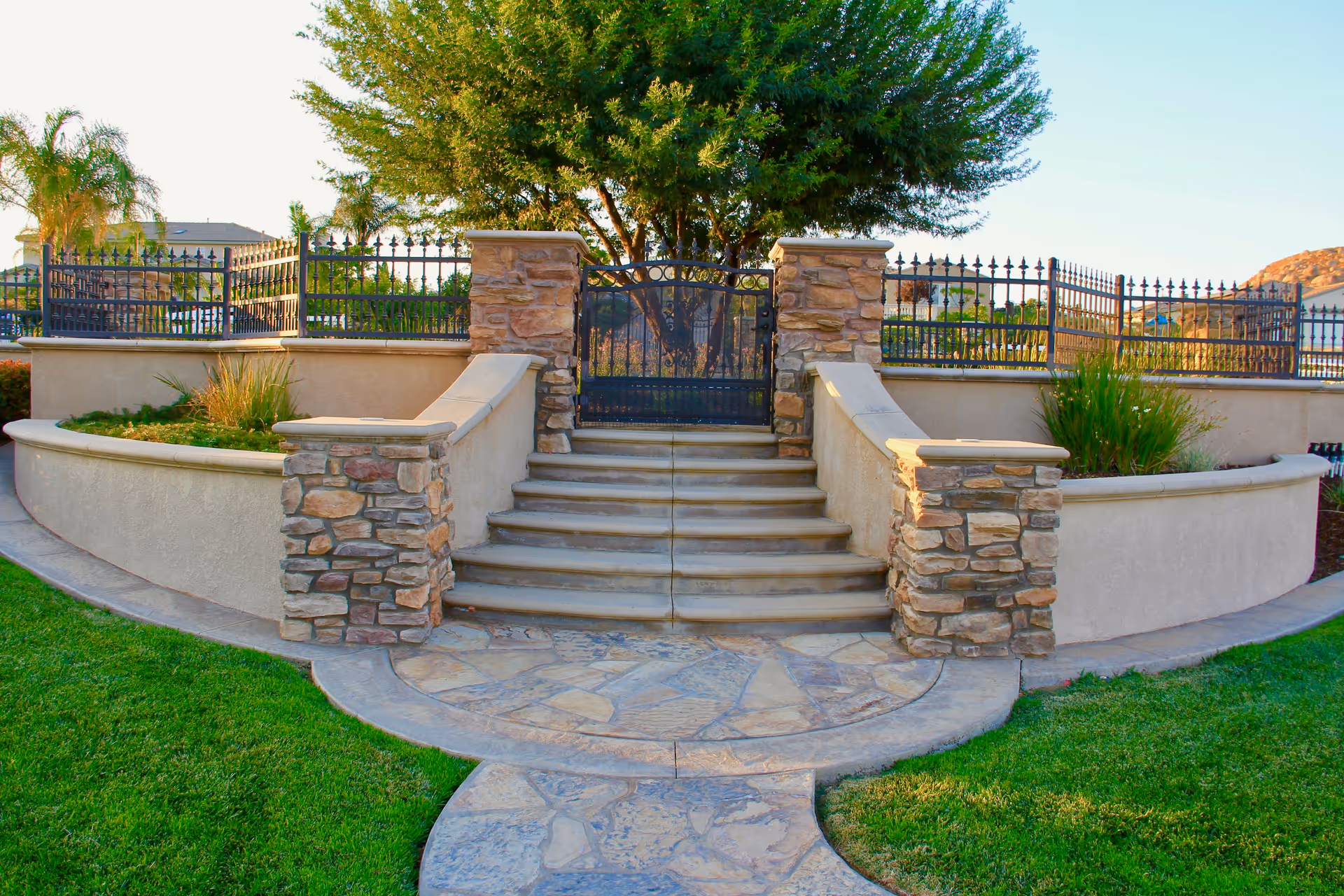 Stone steps leading up to a black wrought iron gate with stone pillars on either side, surrounded by a curved beige wall and green grass lawn, with trees and houses visible in the background under a clear sky.