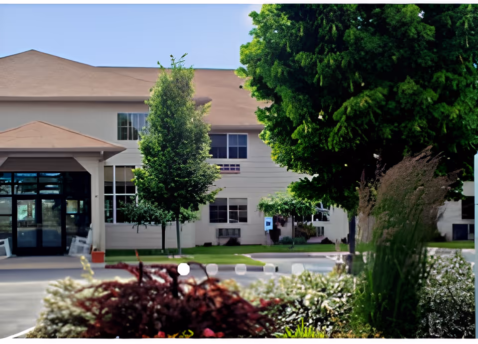 Exterior view of a senior living facility building with a beige facade and brown roof, surrounded by green trees and landscaped bushes in the foreground.