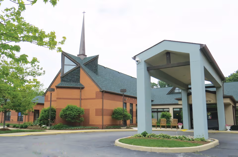 Exterior view of Genacross Lutheran Services-Wolf Creek Campus showing a brick building with a tall steeple topped with a cross, surrounded by greenery and trees. There is a covered entrance with a driveway and some wooden chairs near the building entrance.