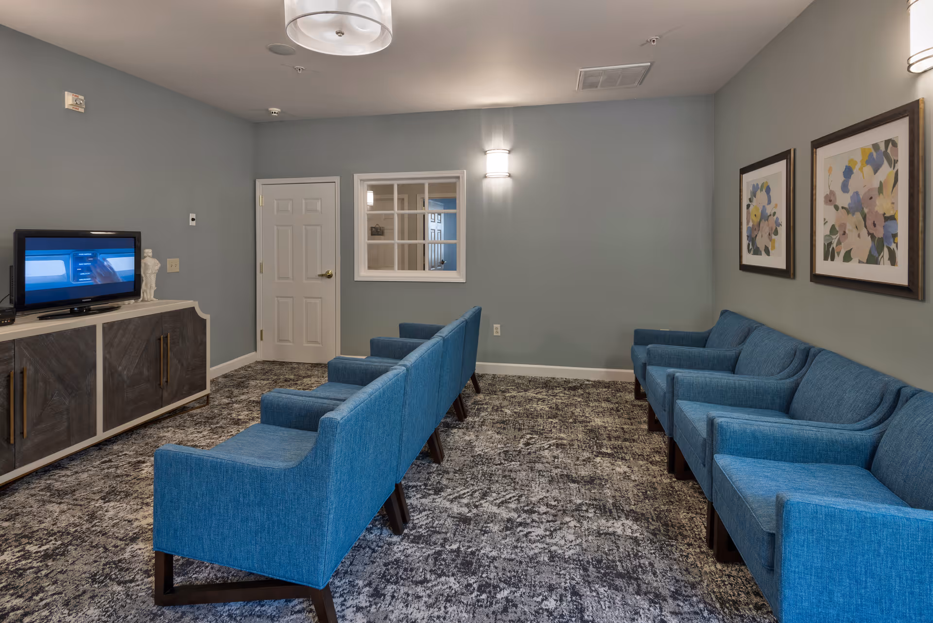 A waiting or common area with two rows of blue upholstered chairs facing a TV on a wooden cabinet. The room has light gray walls, a patterned carpet, two framed floral paintings on the right wall, a white door, and a window with white trim on the back wall. Ceiling lights provide illumination.