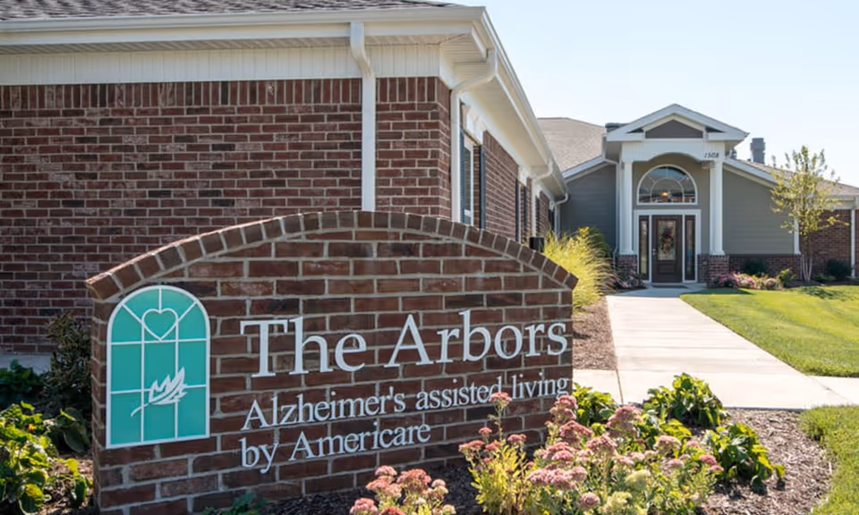 Brick entrance sign for 'The Arbors Alzheimer's assisted living by Americare' with landscaped beds and the facility's front entrance in the background.