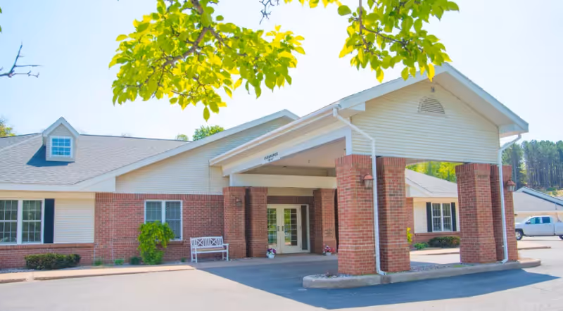 Exterior view of Brookridge Heights facility showing a single-story building with a covered entrance supported by brick columns, white siding, and a bench near the entrance. There are some trees and shrubs around the building and a white vehicle parked nearby.