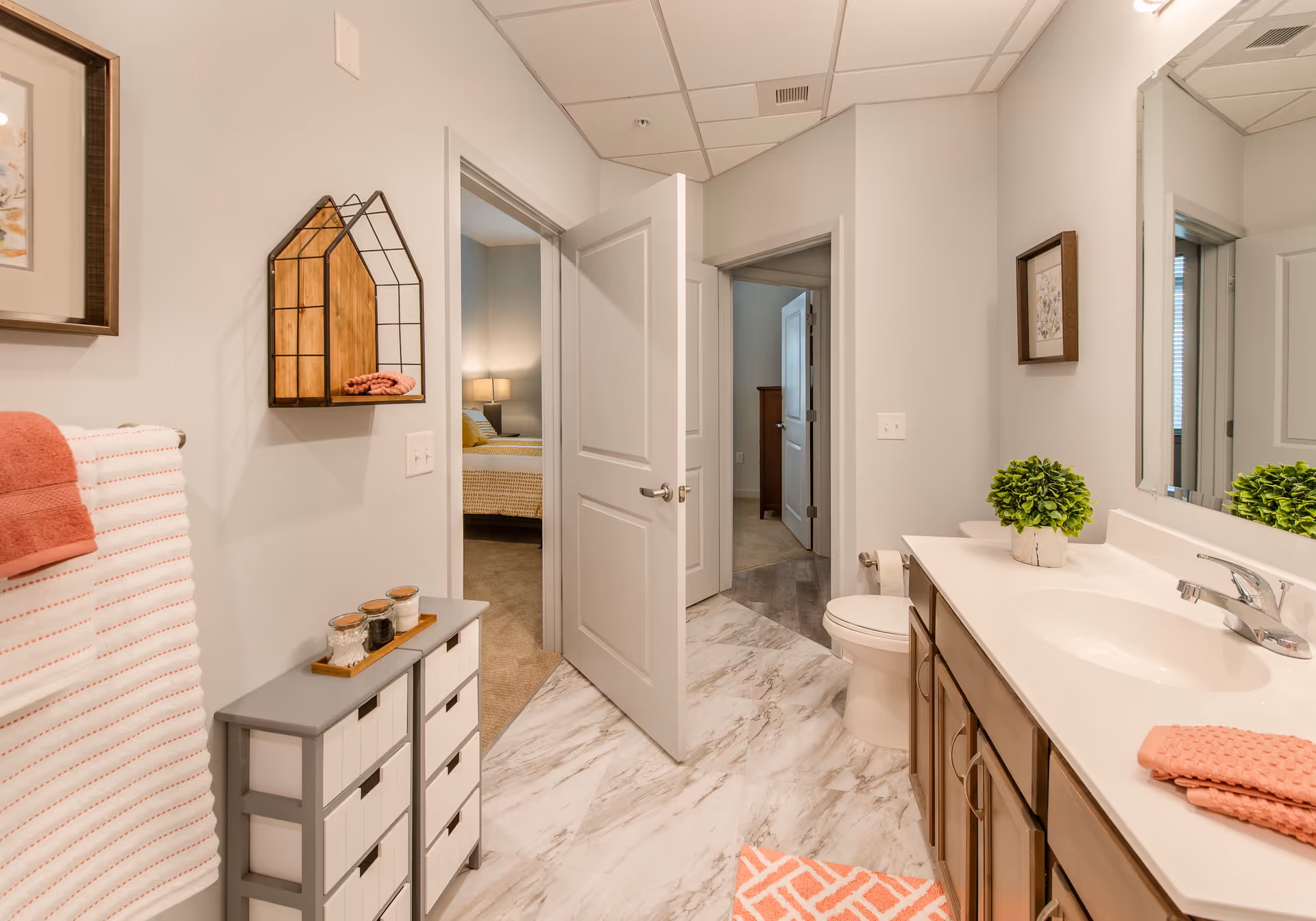 A clean and modern bathroom with a white countertop sink, wooden cabinets, a toilet, and a large mirror. The floor has a marble-like pattern, and there are pink and white towels on a rack and countertop. The bathroom opens into a hallway with doors leading to other rooms, including a bedroom with a bed and lamp visible.
