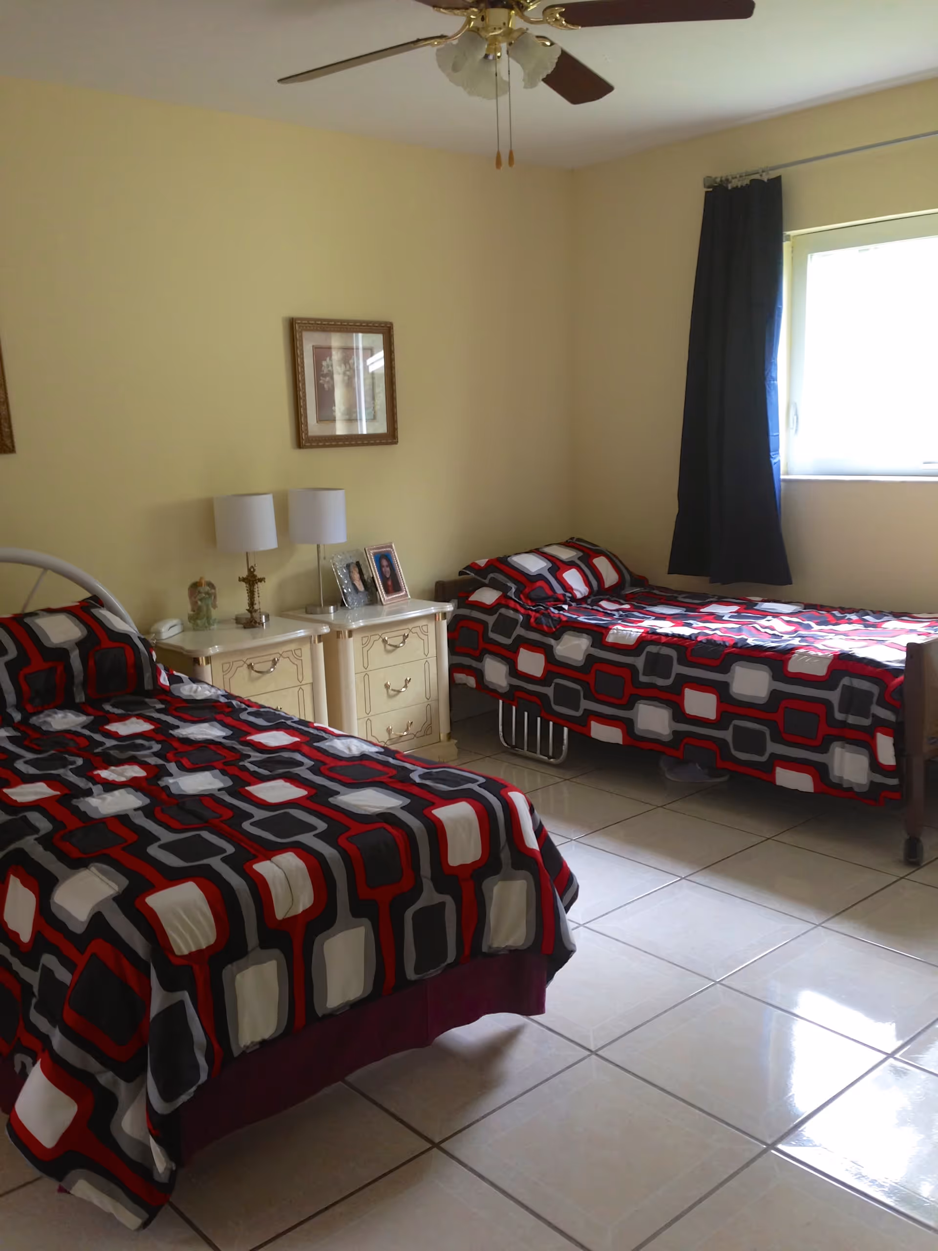 A bedroom with two single beds covered in matching geometric patterned bedspreads in red, black, gray, and white. Between the beds is a cream-colored nightstand with two lamps, framed photos, and a small decorative item. The room has beige walls, a ceiling fan with lights, a window with a dark curtain, and tiled flooring.