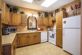 A kitchen with wooden cabinets, beige countertops, a white refrigerator, white stove, white dishwasher, microwave, and various decorative plants and items on top of the cabinets. The floor is tiled and the walls are painted a warm beige color.