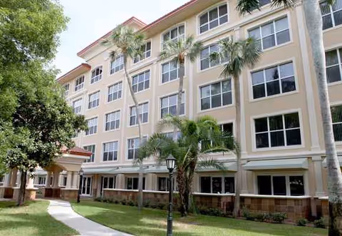 Three-story beige senior living building with palm trees and a walkway courtyard.