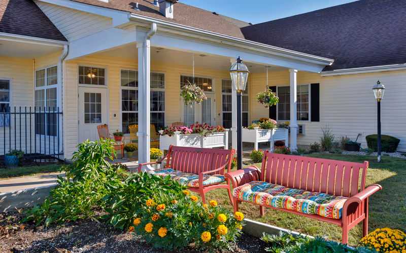 Outdoor seating area at 1019 Belle's Place of Crawfordsville featuring two red benches with colorful cushions, surrounded by flower beds and hanging flower baskets under a covered porch attached to a light yellow building.