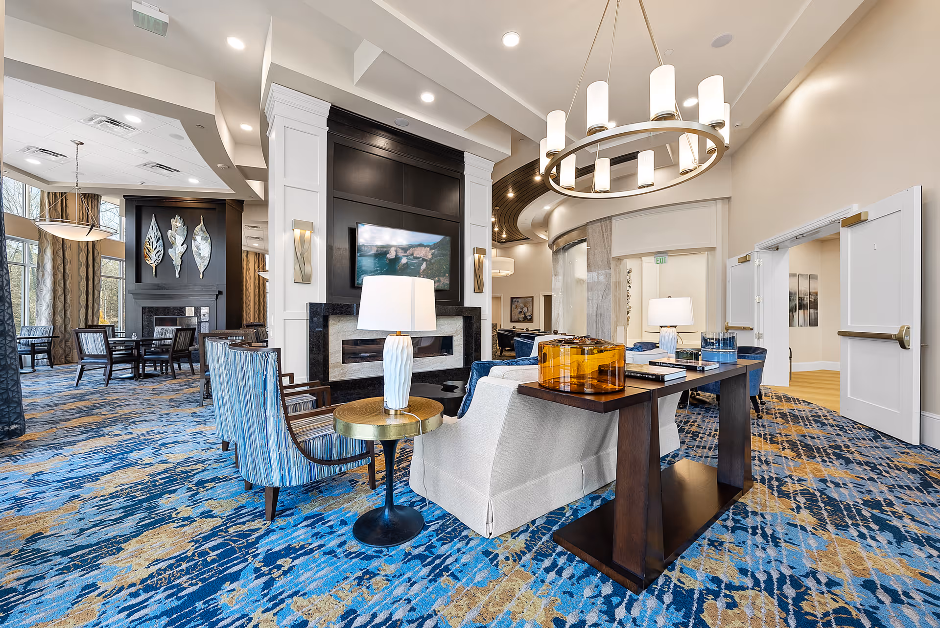 A spacious and well-lit living room area in a senior living facility featuring a modern fireplace with a mounted TV above it. The room has comfortable seating including armchairs and sofas arranged around a dark wood console table with decorative items. The carpet has a blue and gold pattern, and large windows with curtains allow natural light to fill the space. There are also dining tables and chairs visible in the background.