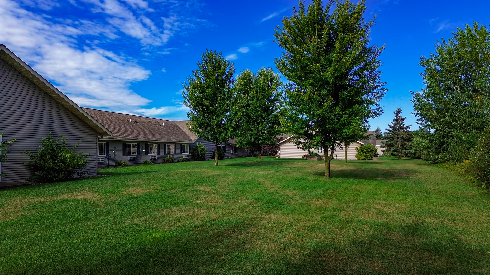 A grassy outdoor area with several green trees and a clear blue sky with some clouds. There are single-story buildings with gray siding and multiple windows surrounding the lawn.