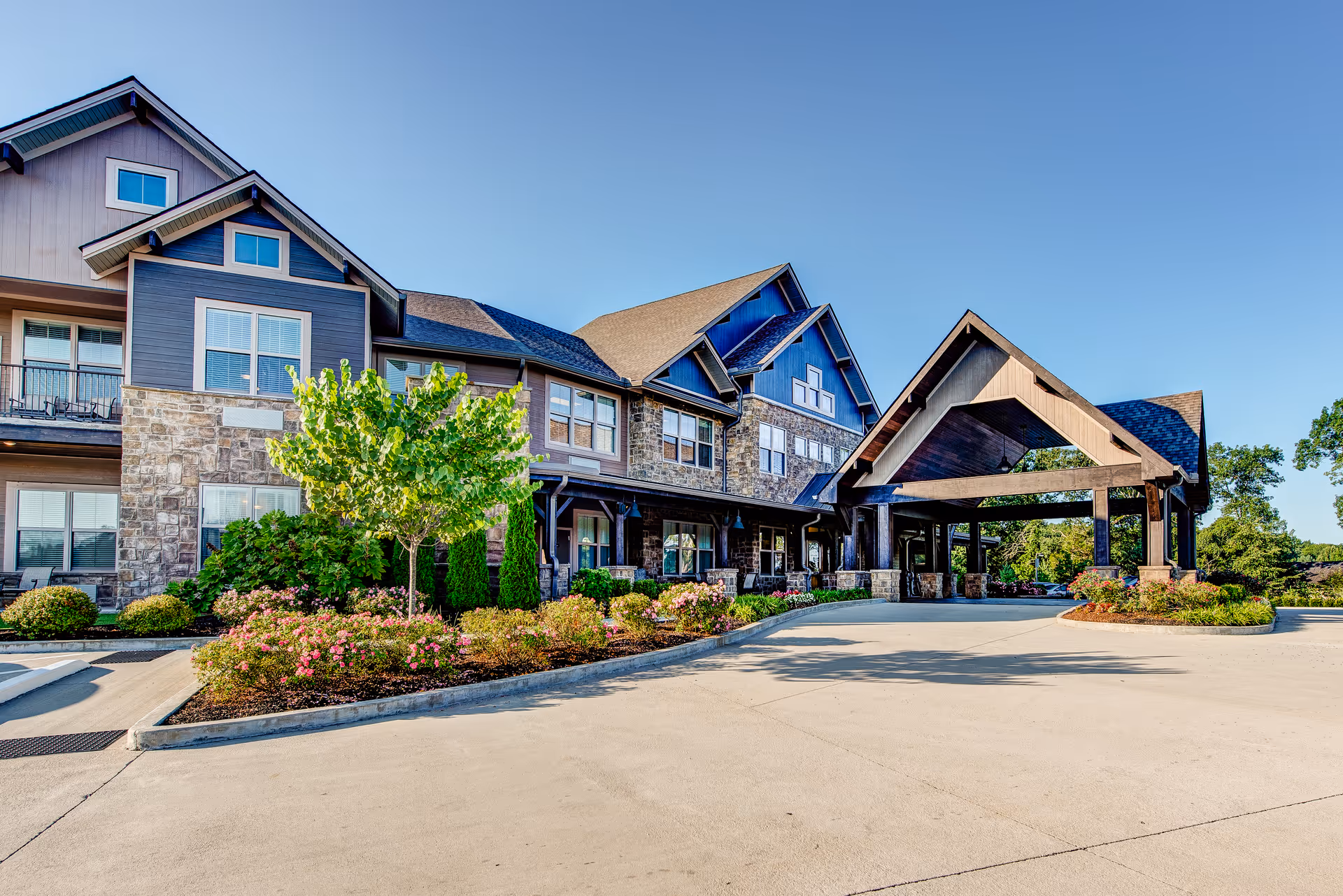 Large two-story senior living building with stone and wood siding, landscaped entrance and covered porte-cochère under a clear blue sky.