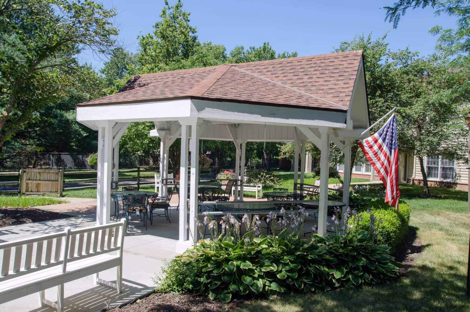 White gazebo pavilion with outdoor tables and chairs, an American flag, and surrounding landscaping in a courtyard.