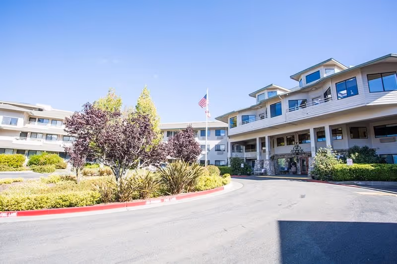 Exterior view of a multi-story retirement community building with a circular driveway, landscaped greenery including trees and bushes, and an American flag on a flagpole in front of the entrance under a clear blue sky.