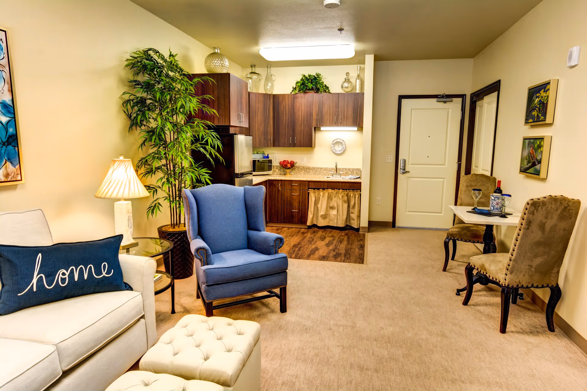 A cozy living area in a senior living facility featuring a white sofa with a blue pillow that says 'home', a blue upholstered armchair, a small round glass side table with a lamp, a potted plant, and a small kitchen area with wooden cabinets, a sink, and a refrigerator. There is also a small dining table with two upholstered chairs and wall art near the entrance door.