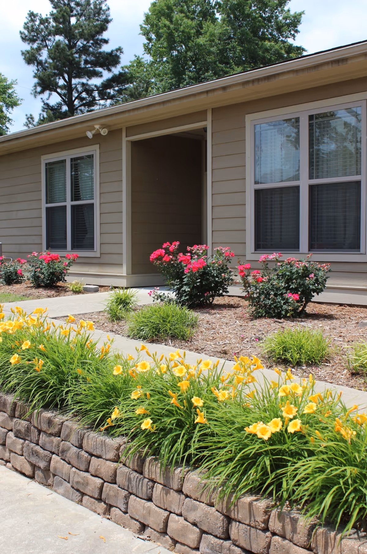 Exterior view of a senior living facility building with beige siding, two large windows, and a small covered entrance. In front of the building, there is a landscaped garden with yellow and pink flowers, green shrubs, and a stone retaining wall.