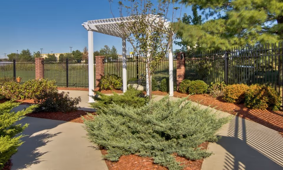 Outdoor garden area with a concrete pathway, green shrubs, small trees, and a white pergola structure. The area is enclosed by a black metal fence with brick pillars, and there is a clear blue sky above.