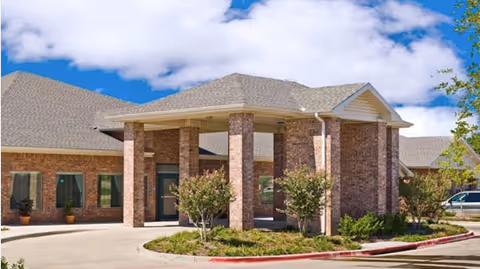 Front entrance of a brick senior living building with a covered porte-cochère, columns, landscaping and driveway under a blue sky.