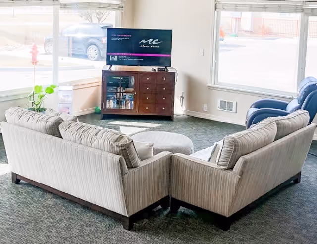 A bright living room area with two beige striped sofas arranged in an L-shape around a round ottoman. A flat-screen TV is placed on a wooden cabinet against the wall, with large windows letting in natural light and showing a view of parked cars outside.