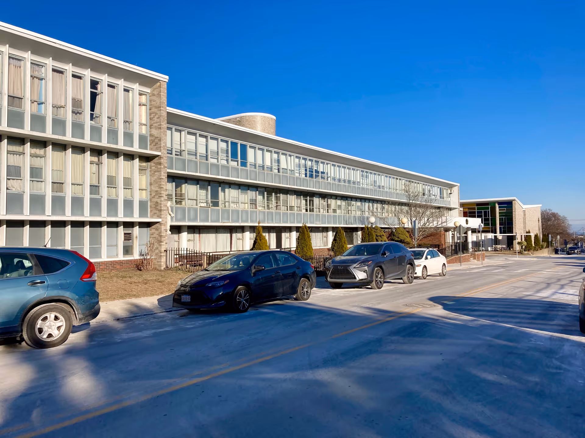 Three-story senior living facility building facade with parked cars along the street under a clear blue sky.