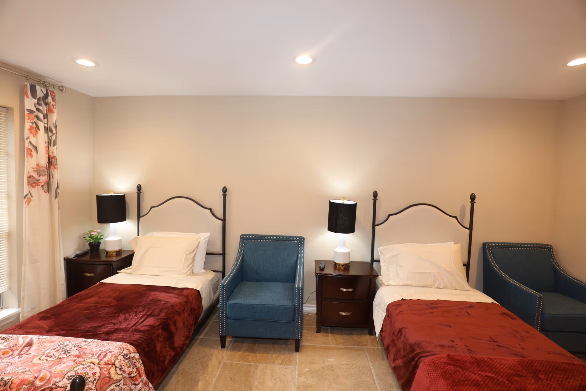 A bedroom with two single beds each covered with a red blanket and white pillows. Between the beds is a dark wooden nightstand with a white lamp and a remote control. Two blue upholstered armchairs are placed next to each bed. The room has beige walls, tiled floor, and a window with floral curtains on the left side.