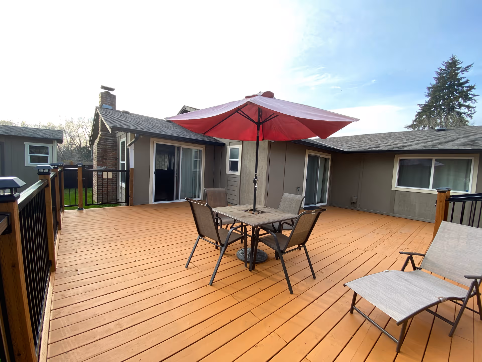 A spacious outdoor wooden deck with a square table, four chairs, and a large red umbrella in the center. There is also a lounge chair on the right side. The deck is surrounded by a railing and is attached to a building with multiple sliding glass doors and windows. Trees and a clear sky are visible in the background.