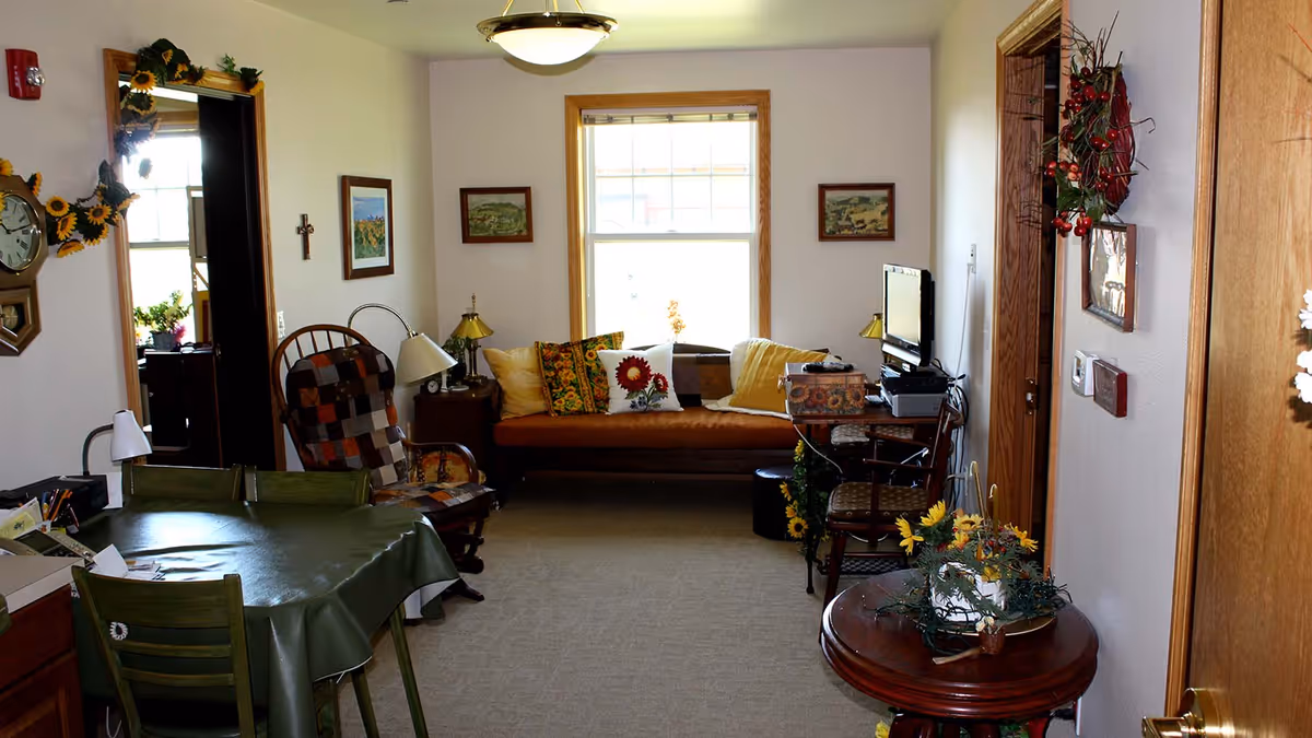 A cozy living room with a brown couch adorned with colorful sunflower-themed pillows, a rocking chair with a patchwork cushion, a small table with a green tablecloth and chairs, and a TV on a stand. The room is decorated with sunflower garlands, framed pictures, and floral arrangements, with a window letting in natural light.