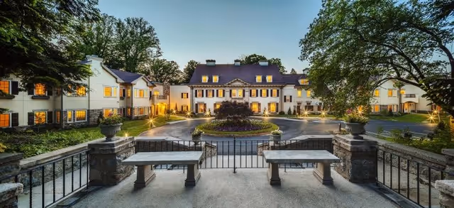 View of a large, elegant senior living facility building at dusk with warm lights glowing from the windows. The building is surrounded by trees and landscaping, with a circular driveway and a central garden area. Stone benches and a wrought iron fence are visible in the foreground.