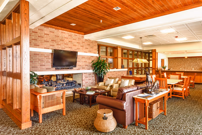 A spacious senior living common area featuring a brick fireplace with a mounted flat-screen TV above it. The room has comfortable seating including leather and fabric armchairs and sofas arranged around a wooden coffee table. There is a wooden partition on the left side, a large potted plant near the fireplace, and a dining area with wooden tables and chairs in the background. The ceiling has recessed lighting and a wooden panel section, and the carpet has a leafy pattern.