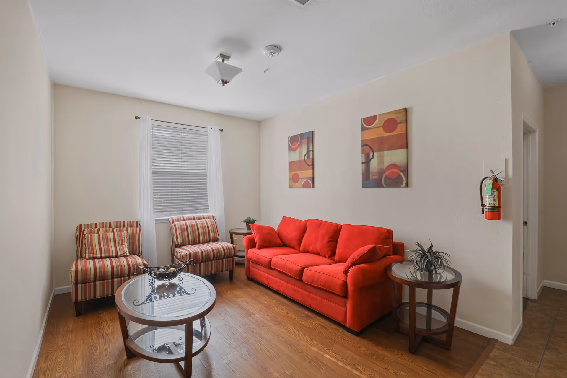 A cozy living room with a bright red sofa, two striped armchairs, a glass coffee table, and a round side table with a plant. Two abstract paintings hang on the beige wall, and a window with white curtains lets in natural light. A fire extinguisher is mounted on the wall near a doorway.