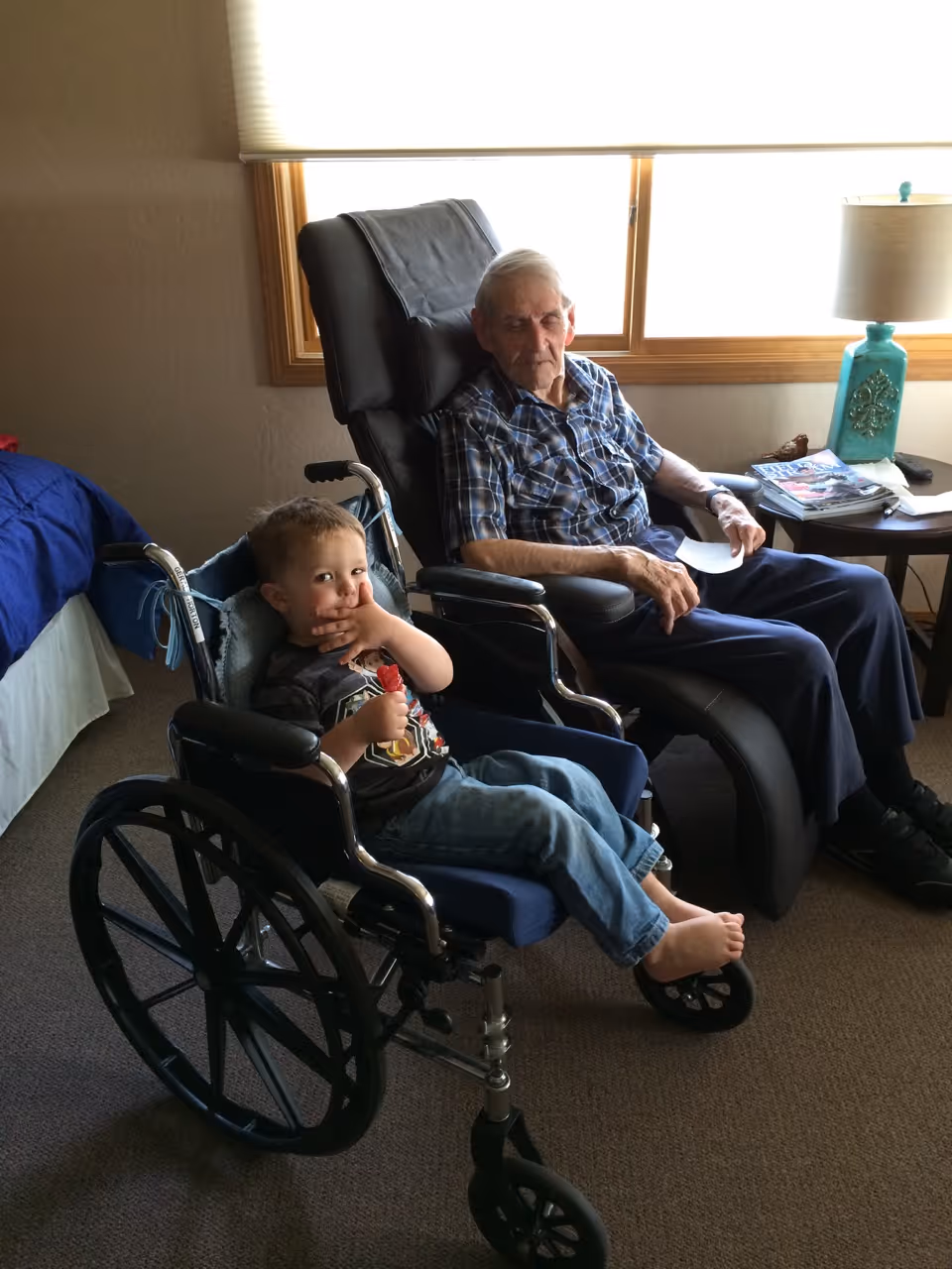 An elderly man sitting in a black recliner chair next to a young boy sitting in a wheelchair inside a room with a bed, a window with blinds, and a side table with a turquoise lamp and magazines.