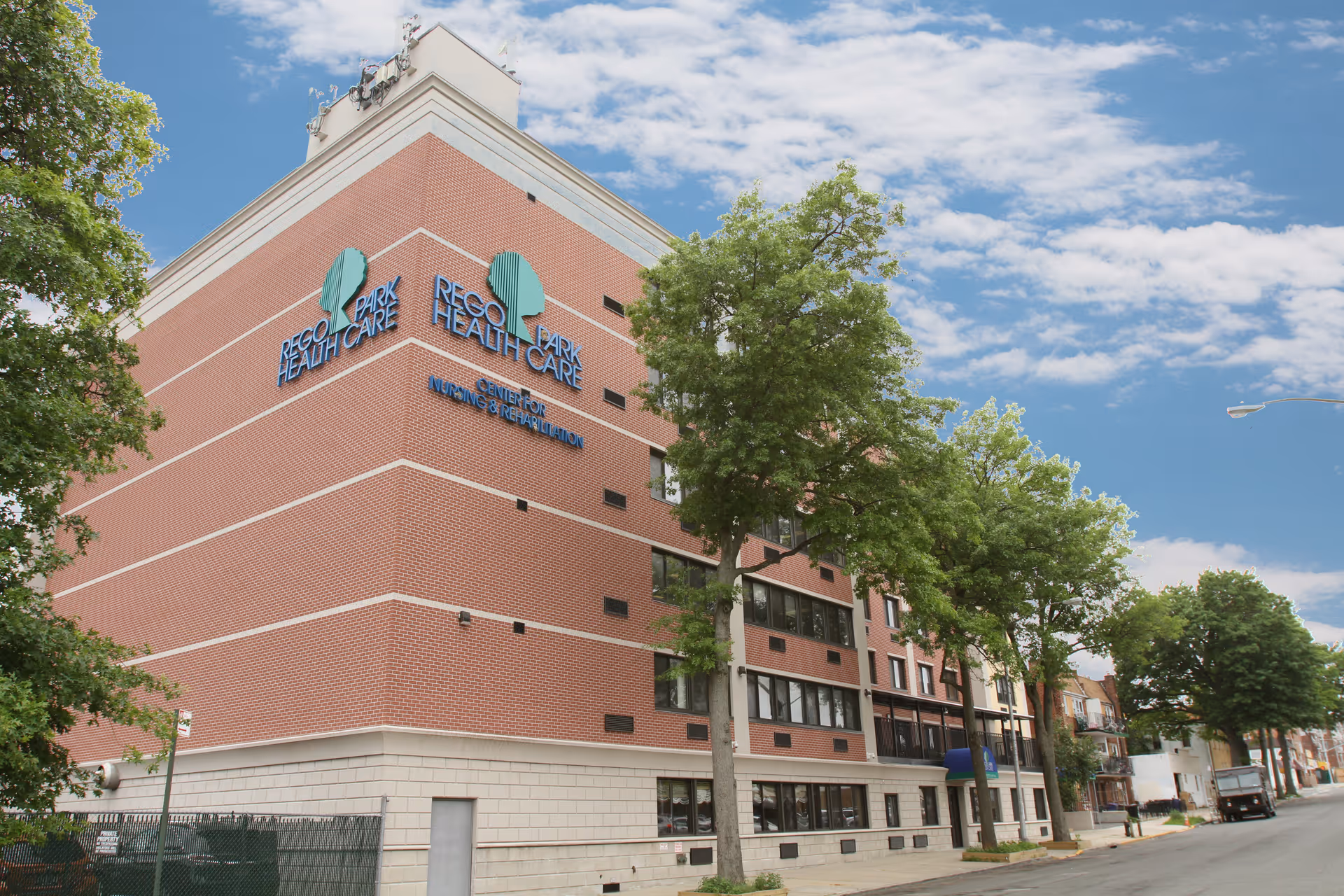 Brick multi-story Rego Park Health Care building on a tree-lined street under a blue sky.