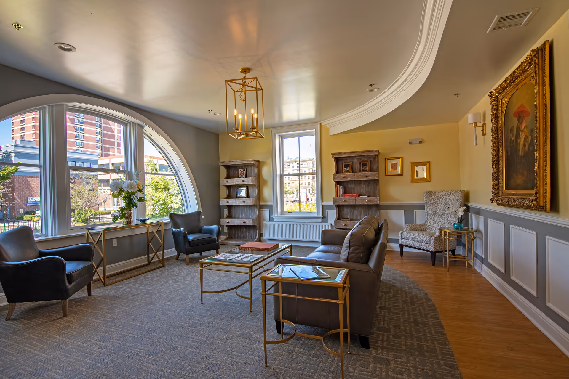 Sunlit common living area with sofas, armchairs, coffee tables, bookshelves and a large arched window overlooking the street.