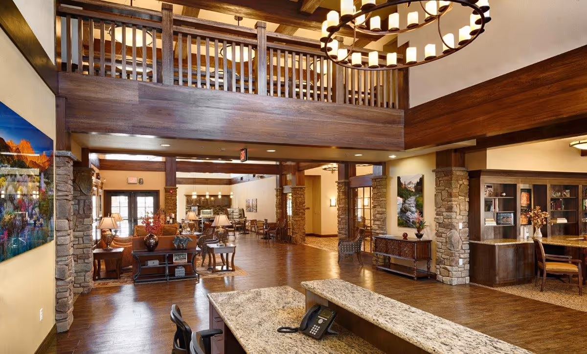 Spacious and warmly lit interior of an assisted living facility featuring a reception desk with a phone, wooden furniture, stone pillars, decorative lamps, and artwork on the walls. The area includes a seating lounge and a dining area in the background, with a second-floor balcony overlooking the space.