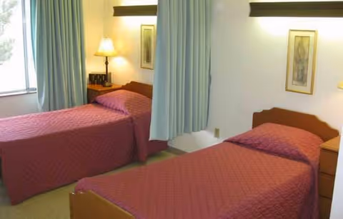A simple bedroom with two single beds covered in matching maroon bedspreads. There is a window with light blue curtains on the left side, a nightstand with a lamp between the beds, and framed artwork on the walls.