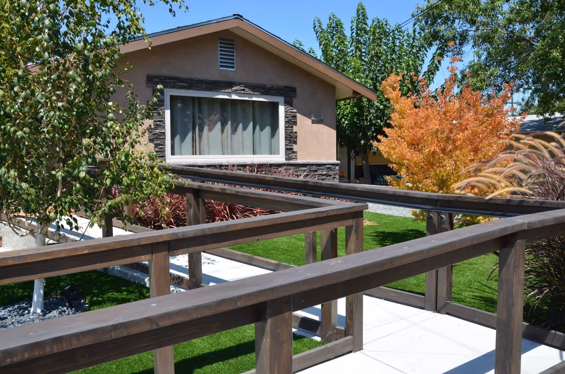 Outdoor view of a single-story building with a large window, surrounded by green grass, trees with green and orange leaves, and a wooden handrail ramp leading to the entrance.