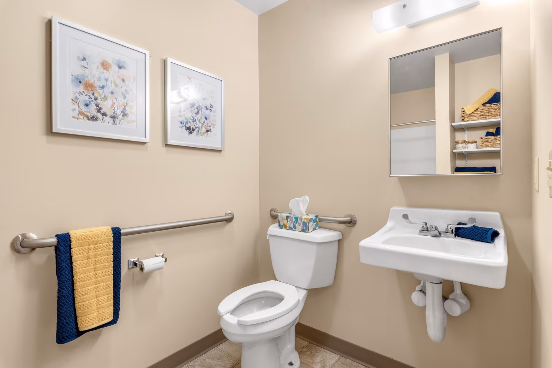 A clean bathroom with beige walls featuring a white toilet and a white sink with a mirror above it. There are two framed floral pictures on the wall, a tissue box on the toilet tank, and a blue and yellow towel hanging on a metal grab bar next to the toilet. The sink has a blue towel placed on its edge, and shelves with baskets and towels are reflected in the mirror.