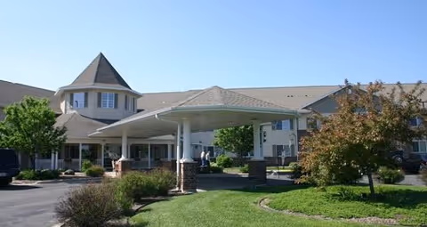 Exterior view of Drumlin Reserve - Assisted Living facility showing a large covered entrance with white pillars and a circular driveway. The building has a turret-like architectural feature and is surrounded by well-maintained landscaping including green grass, bushes, and trees under a clear blue sky.