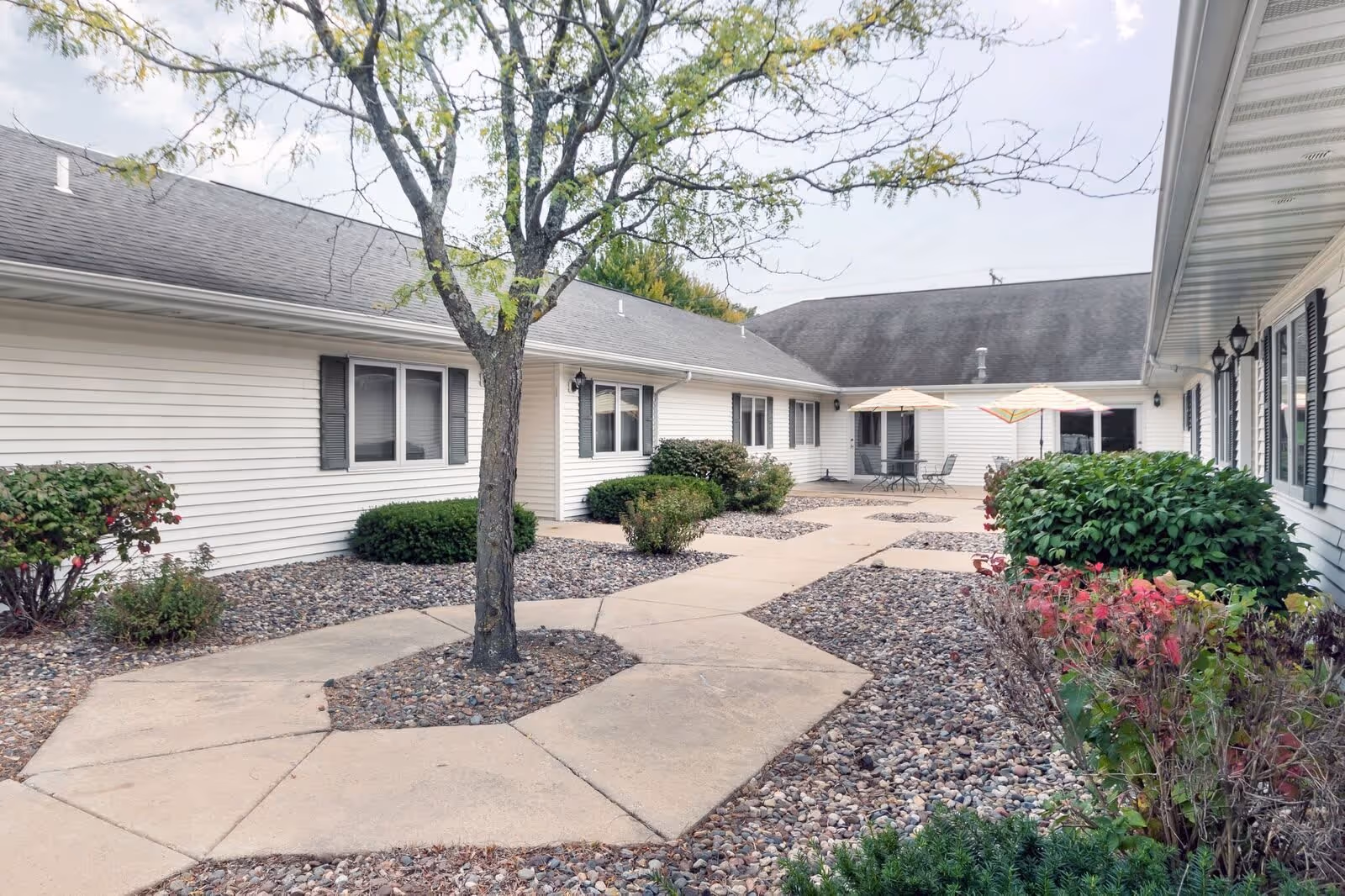 Outdoor courtyard area at Prairie Place Assisted Living with a paved walkway, a tree in the center, bushes, and two patio tables with umbrellas near the building.