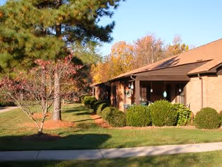 Exterior view of a single-story brick building with a brown roof, surrounded by green bushes and trees with autumn foliage, under a clear blue sky.