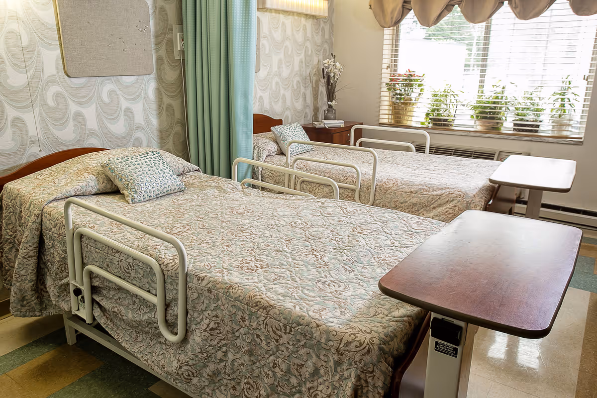 Two twin hospital-style beds with patterned bedspreads, side rails, and overbed tables in a bright shared bedroom with a window and potted plants.