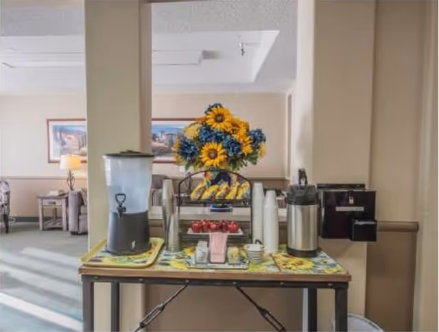 A refreshment table in a common area with a water dispenser, coffee urn, disposable cups, and a tray of bananas and apples. Behind the table is a large bouquet of sunflowers and blue flowers. The background shows a seating area with chairs, a side table with a lamp, and framed artwork on the wall.