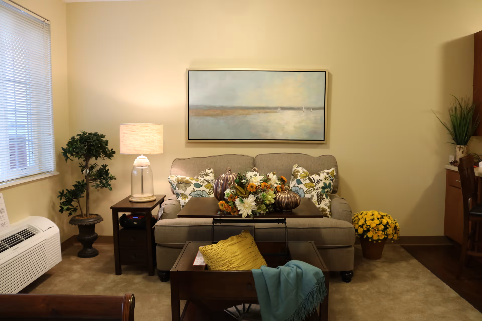 A cozy living room with a beige loveseat adorned with floral pillows. In front of the loveseat is a wooden coffee table with a floral centerpiece and decorative pumpkins. A side table with a lamp and a potted plant is to the left, and a pot of yellow flowers is on the floor to the right. A large framed painting hangs on the wall above the loveseat. The room has beige walls and carpet, with a window covered by blinds on the left side.