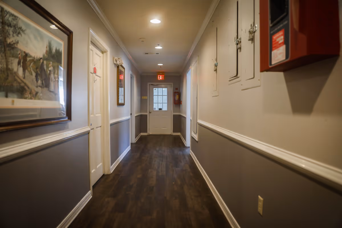 A well-lit hallway in a senior living facility with wood flooring, gray and white walls, several closed doors, a framed painting on the left wall, and an exit door at the end of the corridor. There is a red fire alarm box mounted on the right wall near the foreground.