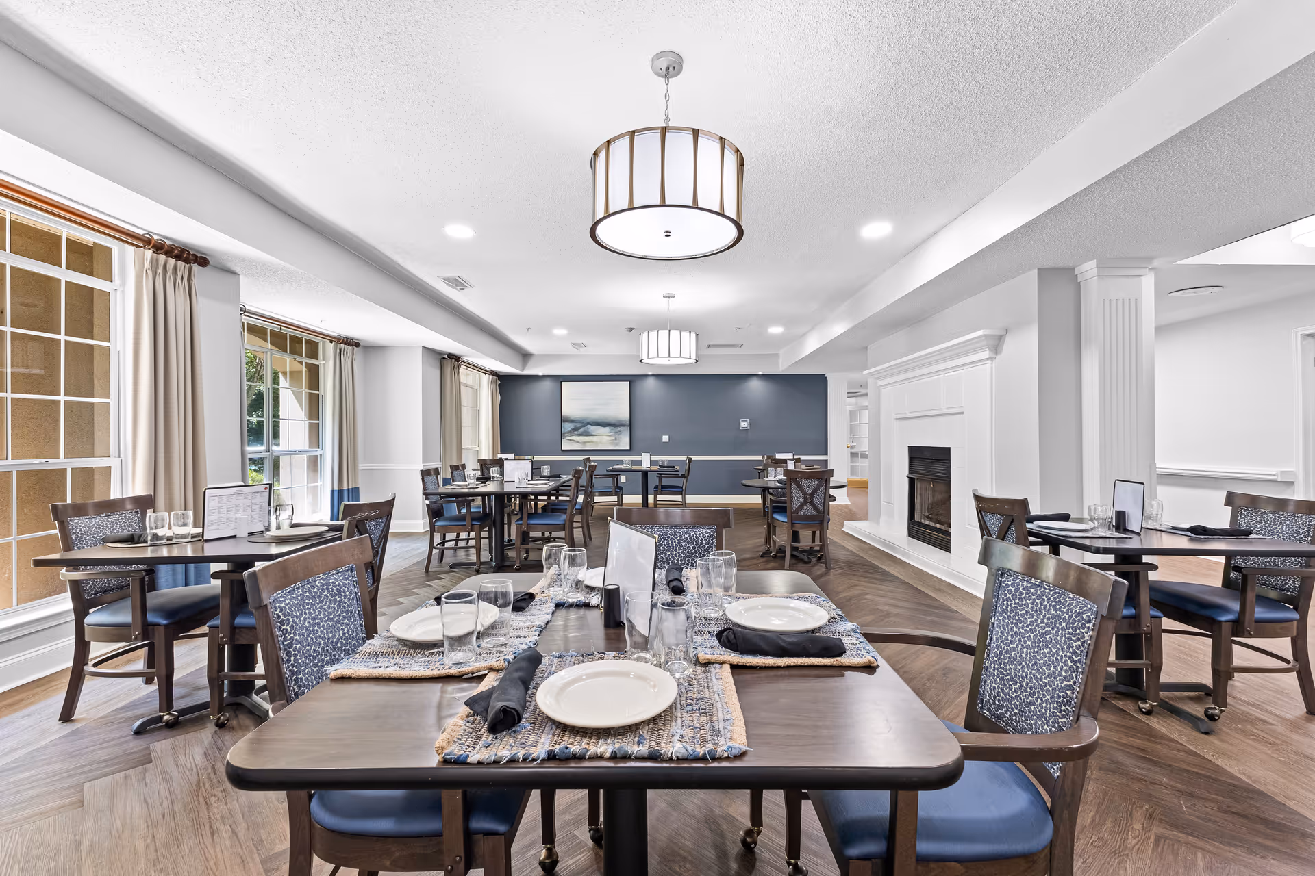 Dining room with multiple set tables and chairs, large windows along one wall, pendant lights, and a fireplace.