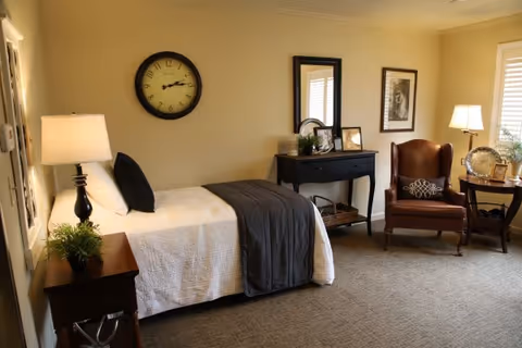 A cozy bedroom in an assisted living community featuring a single bed with white bedding and a dark gray throw blanket, a bedside table with a lamp and a small plant, a wall clock above the bed, a black console table with decorative items and a mirror, a framed picture on the wall, a brown leather armchair with a patterned pillow, a side table with a lamp and decorative items, and a window with white blinds allowing natural light.