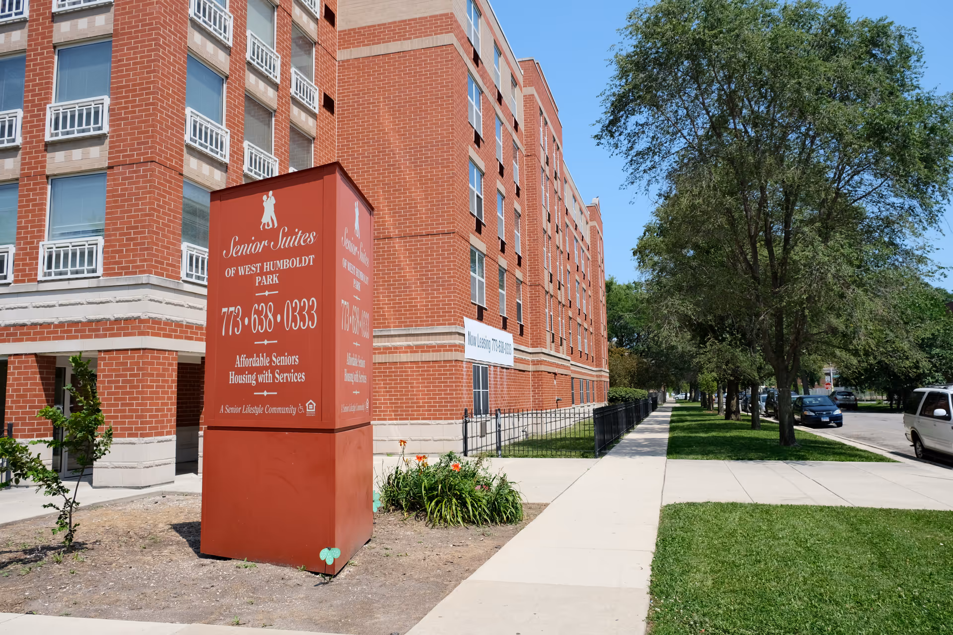 Red-brick Senior Suites of West Humboldt Park building with a large entry sign beside a tree-lined sidewalk.