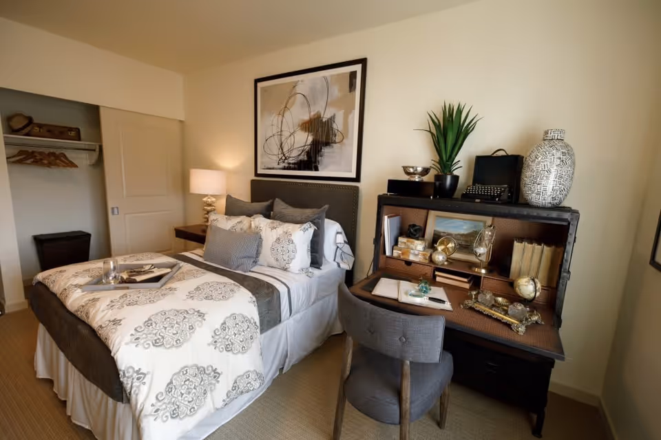 A cozy bedroom featuring a neatly made bed with patterned bedding and multiple pillows. To the right of the bed is a vintage-style desk with an open hutch displaying decorative items, books, and a small globe. A gray upholstered chair is positioned in front of the desk. On the left side of the room, there is an open closet with hangers and a suitcase on the shelf. A bedside table with a lamp is next to the bed, and a large abstract painting hangs above the headboard.