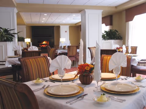 Elegantly set dining room with tables, folded napkins, floral centerpieces and a lit fireplace in the background.