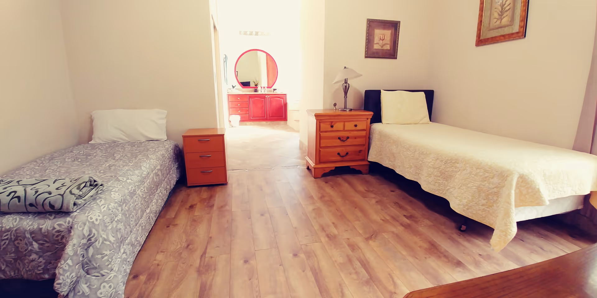 A simple bedroom with two single beds on opposite sides of the room. Each bed has a pillow and a blanket, with a small wooden nightstand between them. The floor is wooden, and the walls are light-colored. There is a doorway leading to a bathroom area with a red vanity and a round mirror. Two framed pictures hang on the walls.