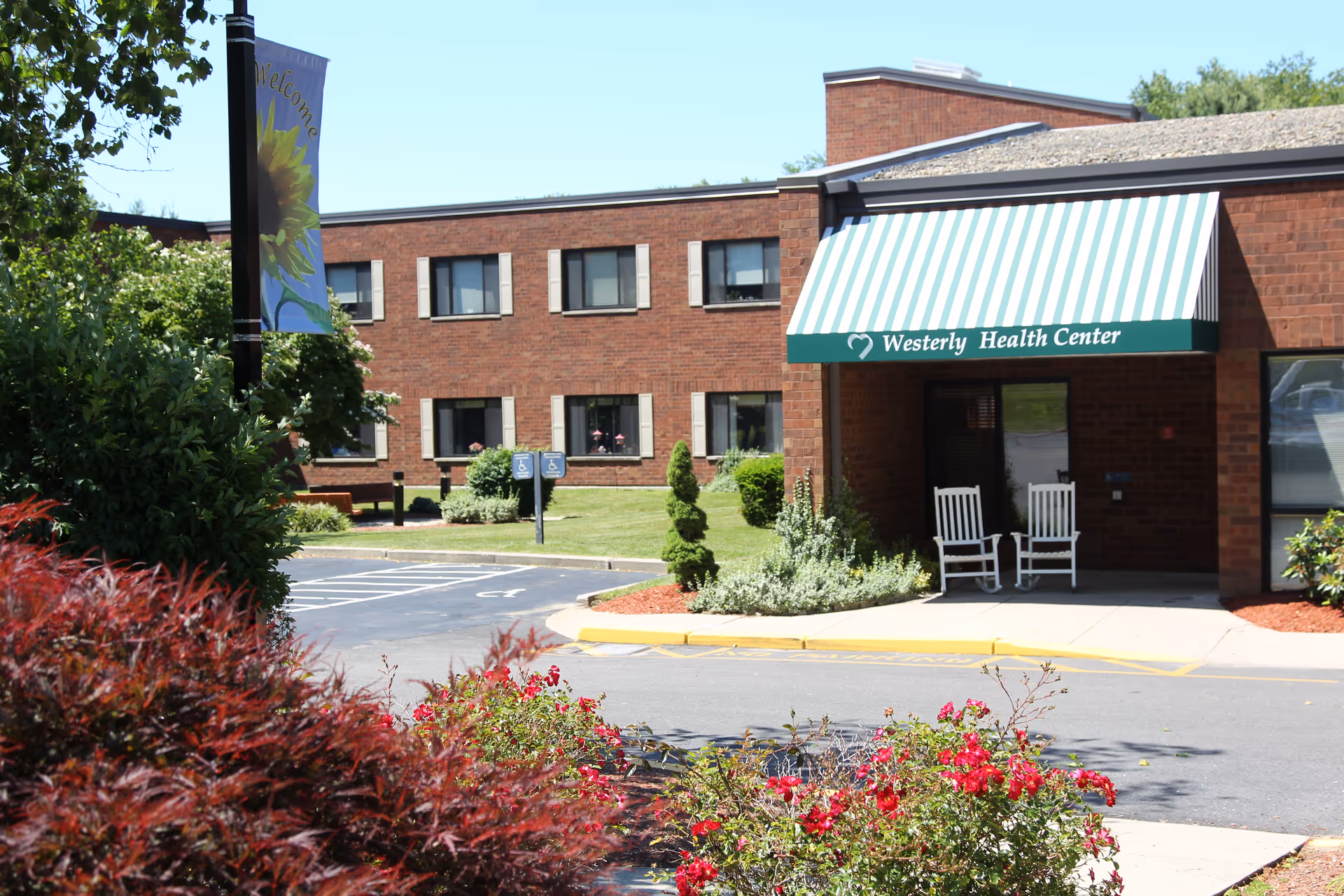 Front entrance of a brick health center with a green-and-white striped awning reading "Westerly Health Center," two white rocking chairs, and landscaped grounds.